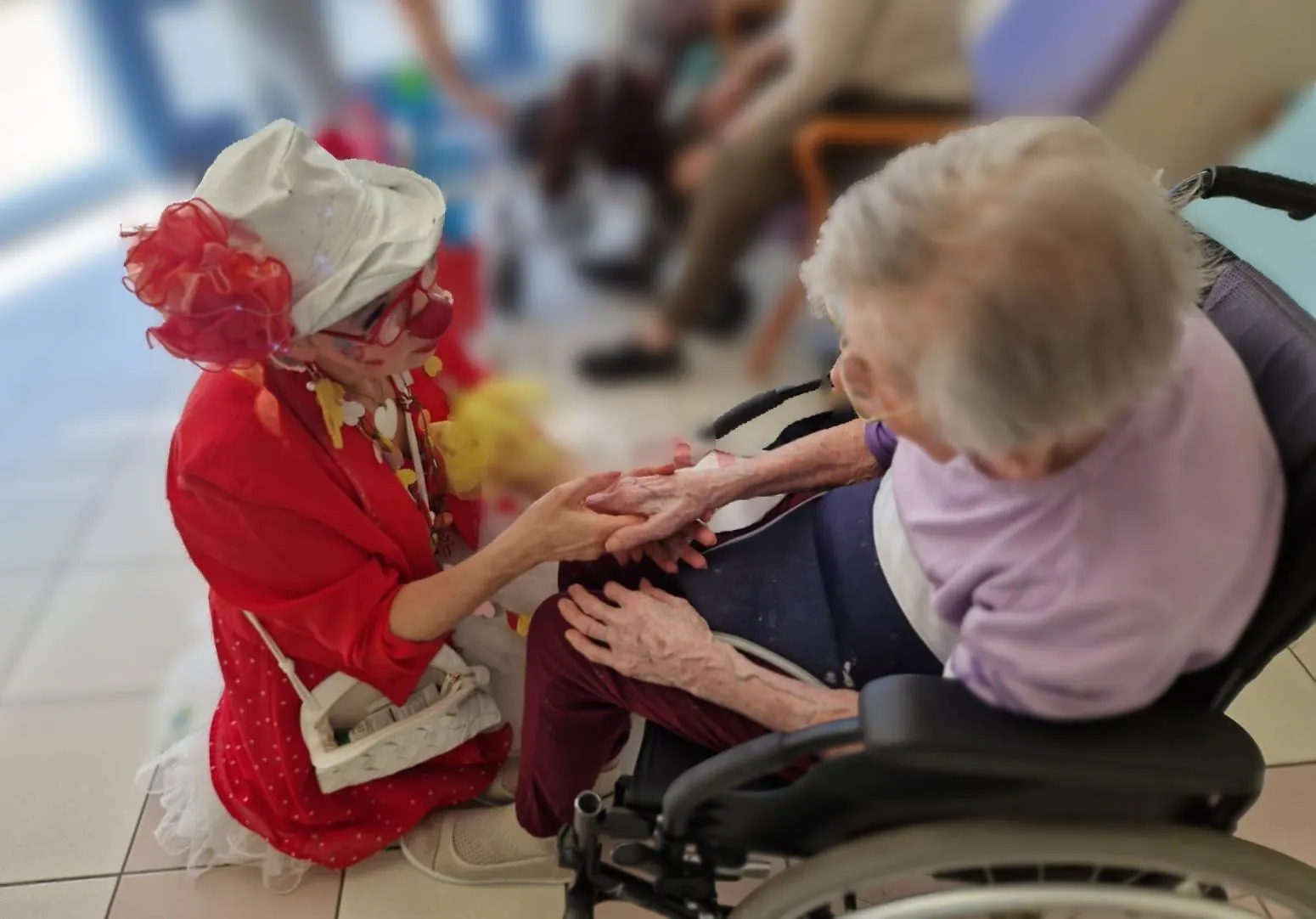 Une clown en tenue rouge, agenouillée à hauteur d'une résidente installée en fauteuil roulant, lui tient les deux mains dans le couloir d'un établissement.