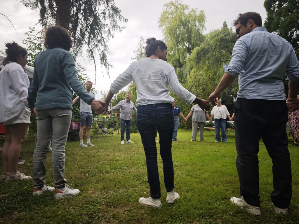 Une dizaine de participants se tiennent par la main, debout en cercle sur une pelouse bordée d'arbres, lors d'une séance en plein air.